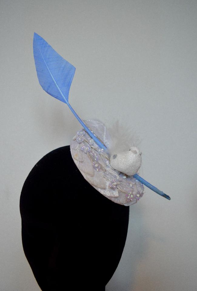 IMAGE - White fascinator covered in velvet leaves and blue mini flowers. Decorated with a white glittered bird and a blue arrow quil.
