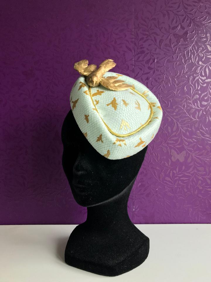 IMAGE - Handblocked fascinator covered in mint fabric, printed with gold birds and a net overlay. Finished with gold ribbon and gold bird. Stays on with an elastic. 