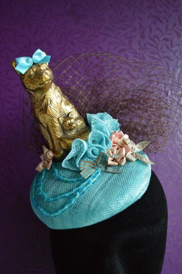 IMAGE - Blue sinamay fascinator with gold cat, brown netting and blue lace. Decorated with blue and brown fabric ornaments and strings of blue beads. Fixes to hair with a comb.