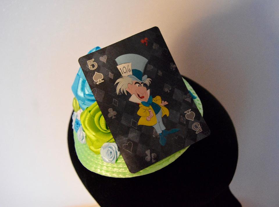 IMAGE - Light green straw fascinator with green and blue ribbon roses and playing card featuring the Mad Hatter. Fixes to hair with a comb.