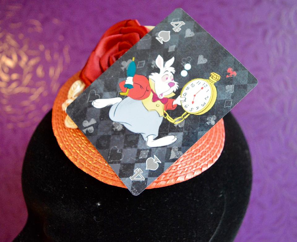 IMAGE - Red straw fascinator, lightly dusted with gold shimmer. Decorated with playing card featuring the White Rabbit and red and cream ribbon roses. Fixes to hair with a comb.